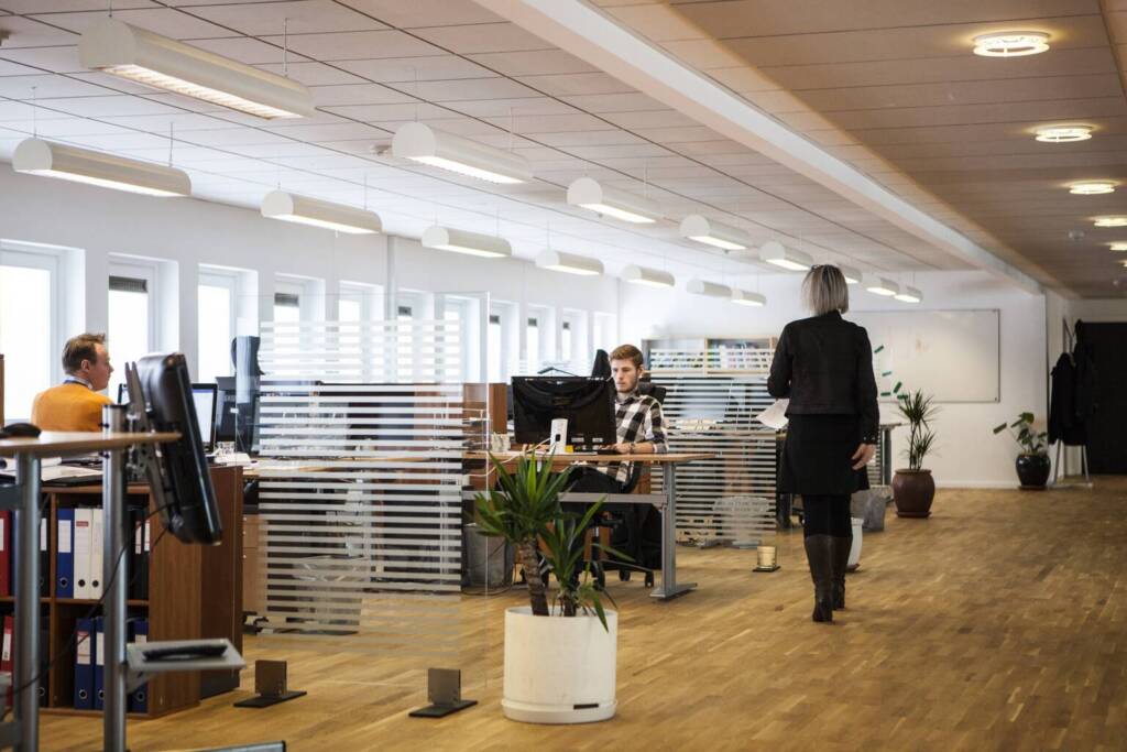 Skills: Government To Invest £1.2 Billion Annually 4 A Modern Office Space With Employees At Their Desks, Computers And Monitors Visible, And A Woman Walking Across The Room. The Setting Includes Overhead Lighting, Frosted Glass Partitions, And Potted Plants.